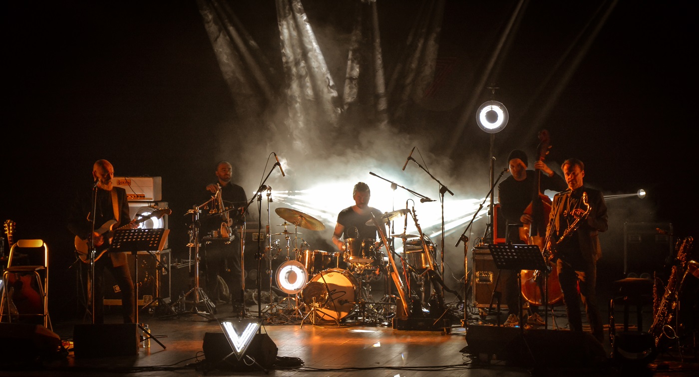 Pięcioosobowy zespół muzyczny na scenie. W centrum perkusista. Po lewej gitarzysta i muzyk grający na niestandardowych instrumentach perkusyjnych. Po prawej kontrabasista i saksofonista. Wszyscy mają czarne stroje. Światła reflektorów rozproszone w sztucznym dymie skupiają się w centrum kadru. Tło po bokach czarne.