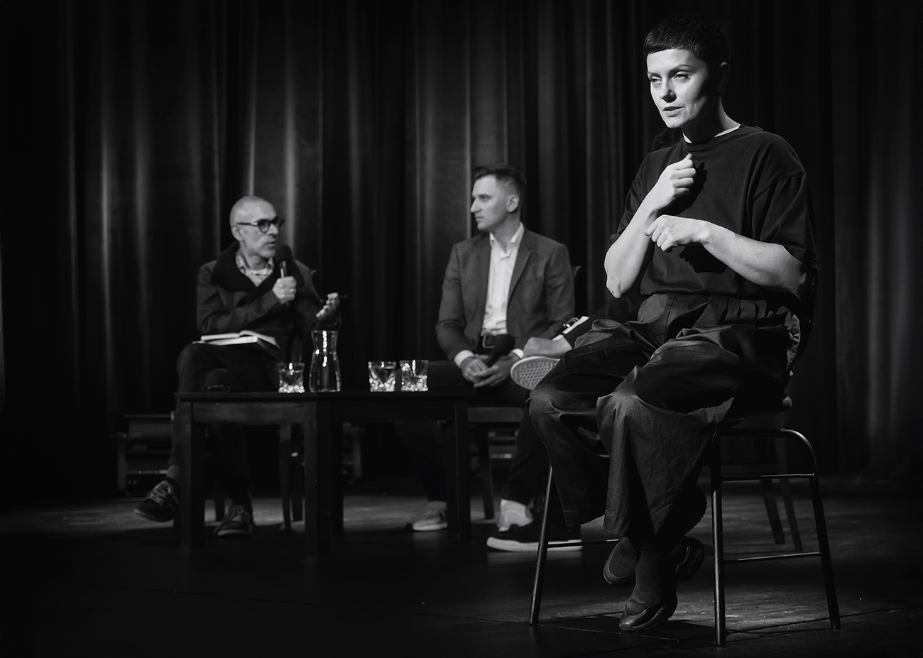 Czarno-białe zdjęcie trzech osób na scenie Teatru. Wszyscy siedzą na krzesłach. Na pierwszym planie po prawej tłumaczka na polski język migowy. Na drugim planie w centrum dwóch mężczyzn mikrofonami przy małym stoliku. W tle ciemna kotara.