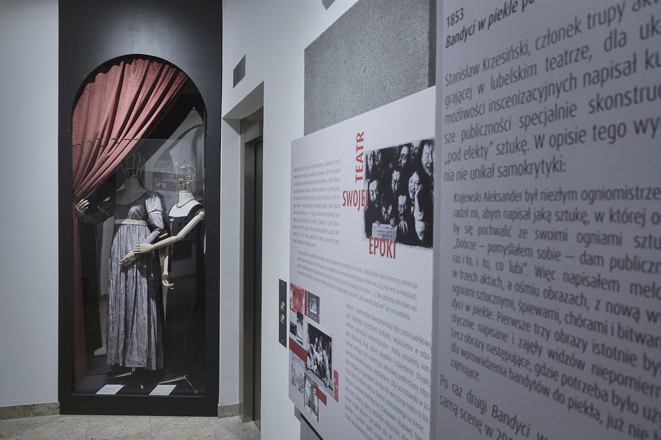 Zdjęcie wystawy wewnątrz Teatru. Po prawej stronie ściana z tablicami z grafikami, zdjęciami i tekstem. W głębi ujęcia po lewej stronie jest gablota z łukiem u góry.