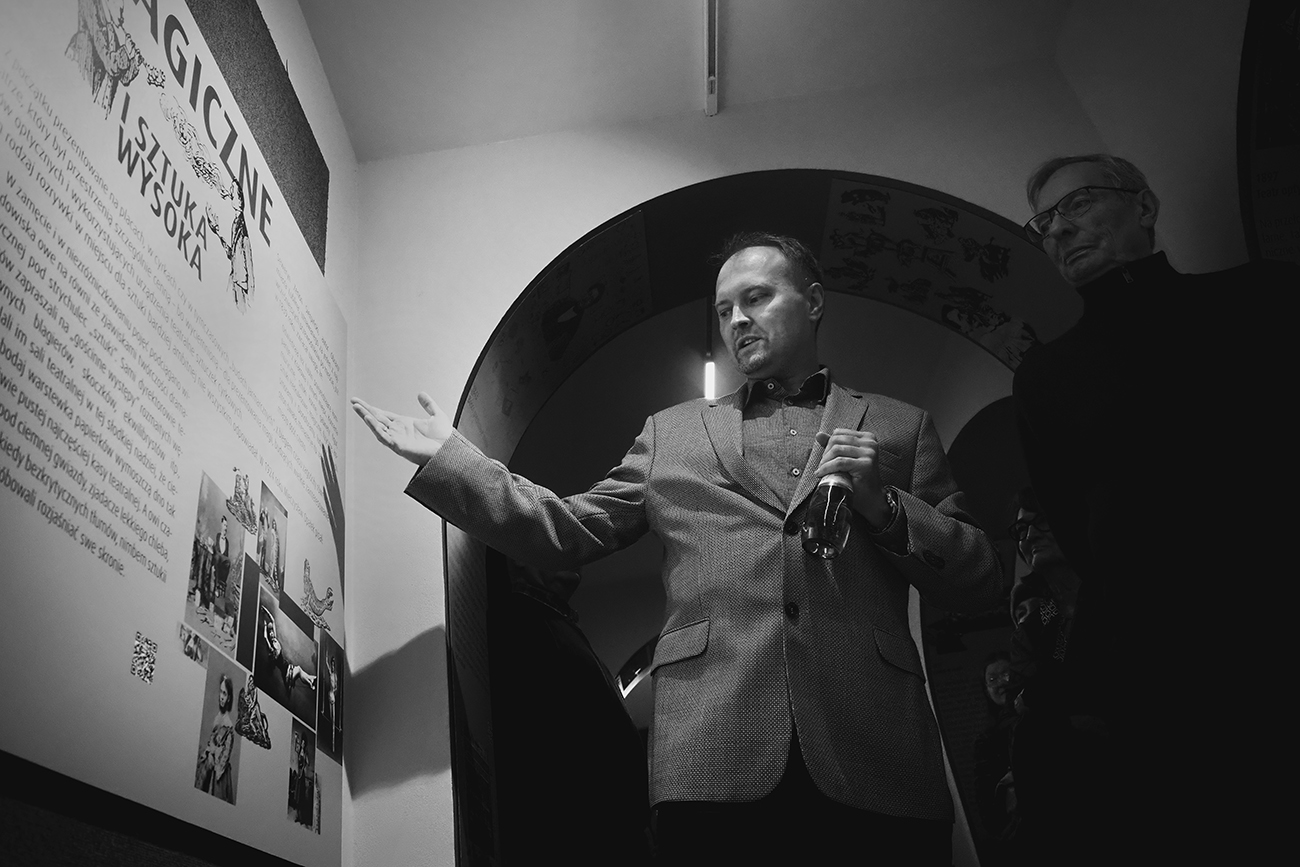 Czarno-białe zdjęcie dwóch mężczyzn we wnętrzu Teatru. Jeden z nich wskazuje dłonią na tablicę na ścianie. Na tablicy są grafiki, zdjęcia i tekst. W tle ścina z wnęką z łukiem, we wnęce jest półmrok.