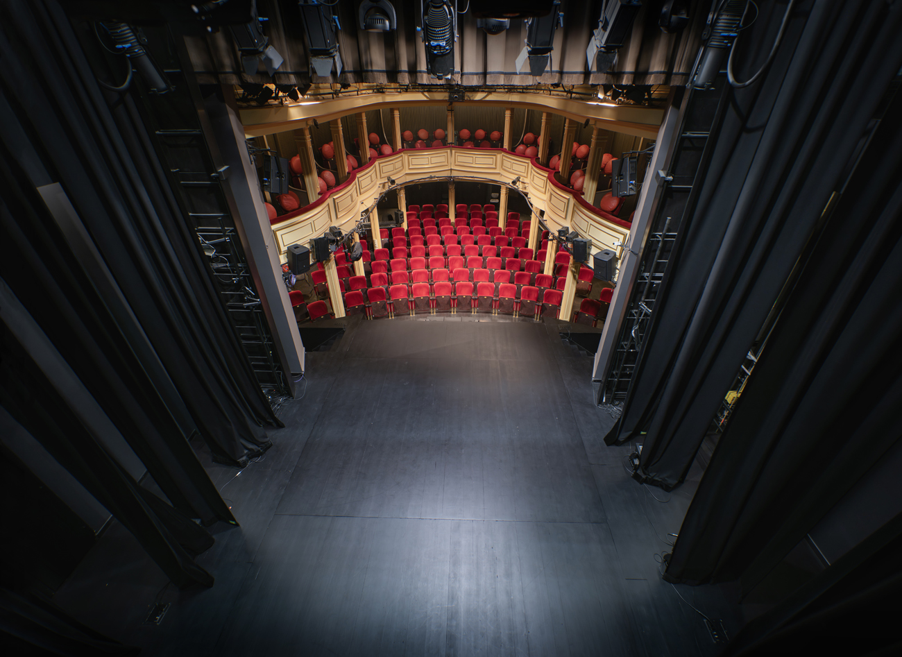 Zdjęcie sceny na sali widowiskowej Teatru Starego. Ujęcie z góry z zascenia. W dolnej części kadru czarna scena. Po bokach aksamitne, wysokie kulisy. W głębi kadru trzypoziomowa widownia z czerwonymi fotelami i kremowymi balustradami na lożach i balkonie, czyli pierwszym i drugim piętrze.