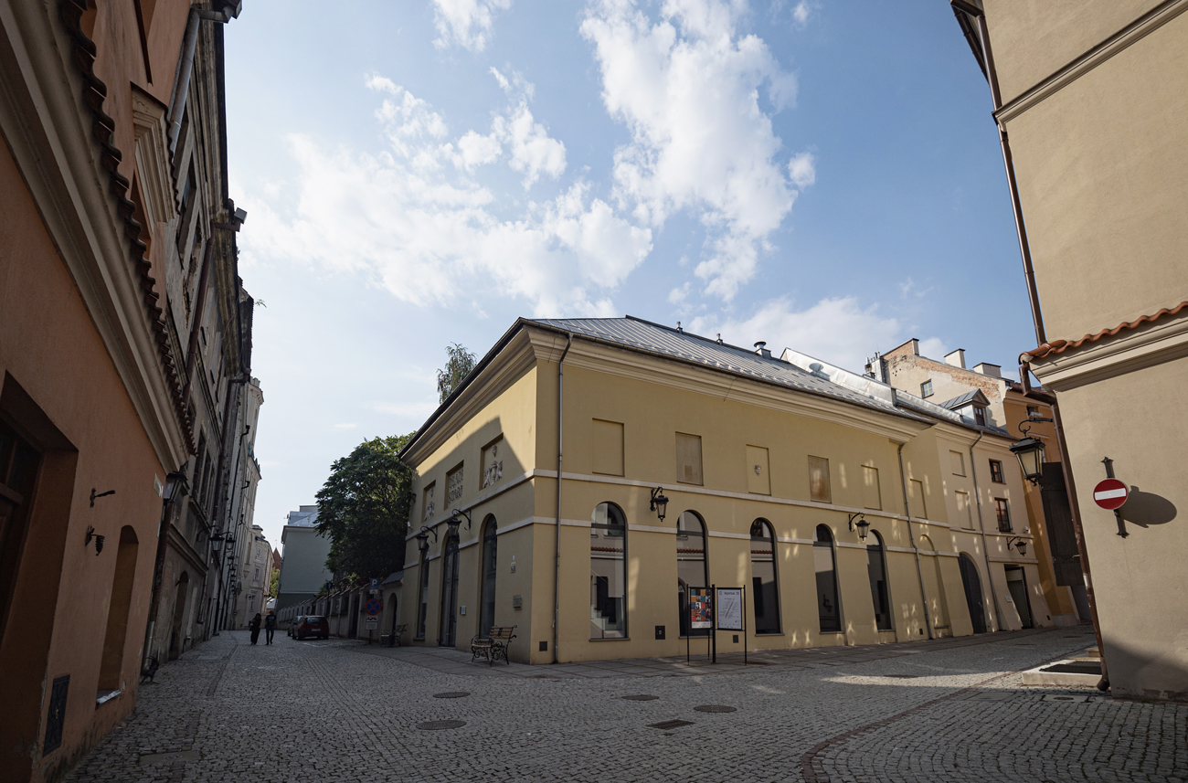 Zdjęcie budynku Teatru Starego w Lublinie i najbliższego otoczenia. Ujęty jest róg budynku. Na prawo fasada od strony ulicy Jezuickiej. Na lewo boczna ściana od strony ulicy Dominikańskiej. Budynek jest bladożółty z białymi gzymsami na wysokości pierwszego piętra. Budynek ma wysokie okna z łukowym zakończeniem. Zaczynają się tuż nad ziemią i sięgają do pierwszego piętra. Nad nimi znajdują się na drugim piętrze płytkie wnęki. Po lewej stronie znajduje się światło ulicy Jezuickiej i rząd kamienic, które przy niej stoją. Po prawej stronie kamienica przy ulicy Dominikańskiej znajdująca się naprzeciwko Teatru. W tle u góry błękitne niebo z drobnymi białymi chmurami.