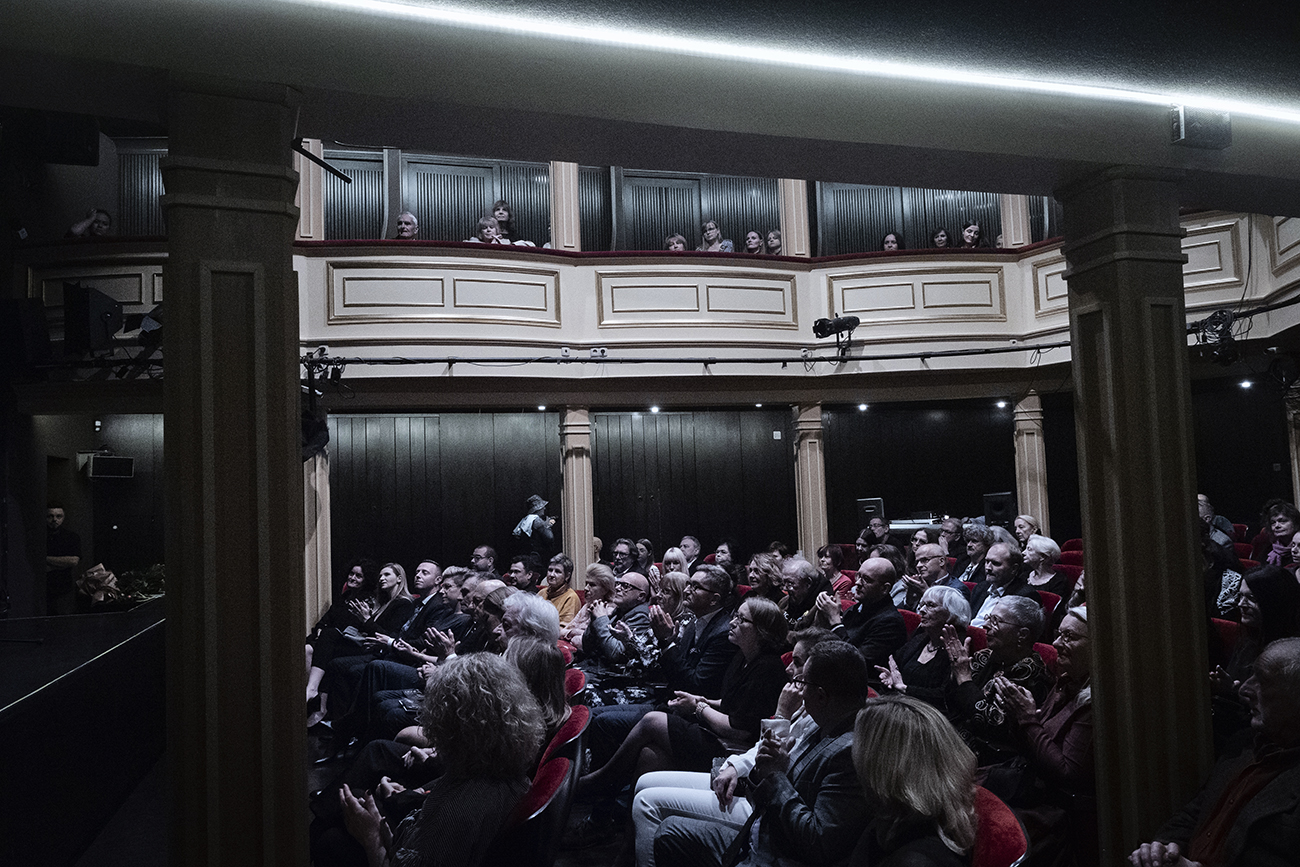 Zdjęcie wykonane wewnątrz sali widowiskowej w Teatrze Starym w Lublinie. Ujęcie z boku sali na parterze. Wszystkie miejsca są zajęte przez widzów. W głębi ujęcia znajdują się loże boczne, wsparte na jasnych kolumnach, mają jasne balustrady. W lożach również są widzowie.