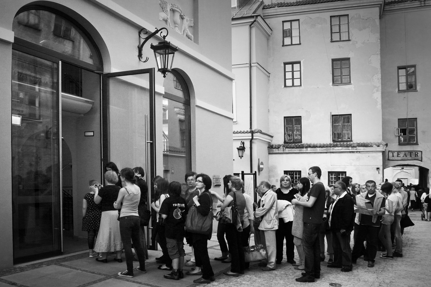 Czarno białe zdjęcie wykonane przed głównym wejściem do Teatru Starego w Lublinie. Szklane drzwi do Teatru są otwarte, a przed nimi ustawiona jest długa kolejka osób czekających na wejście.