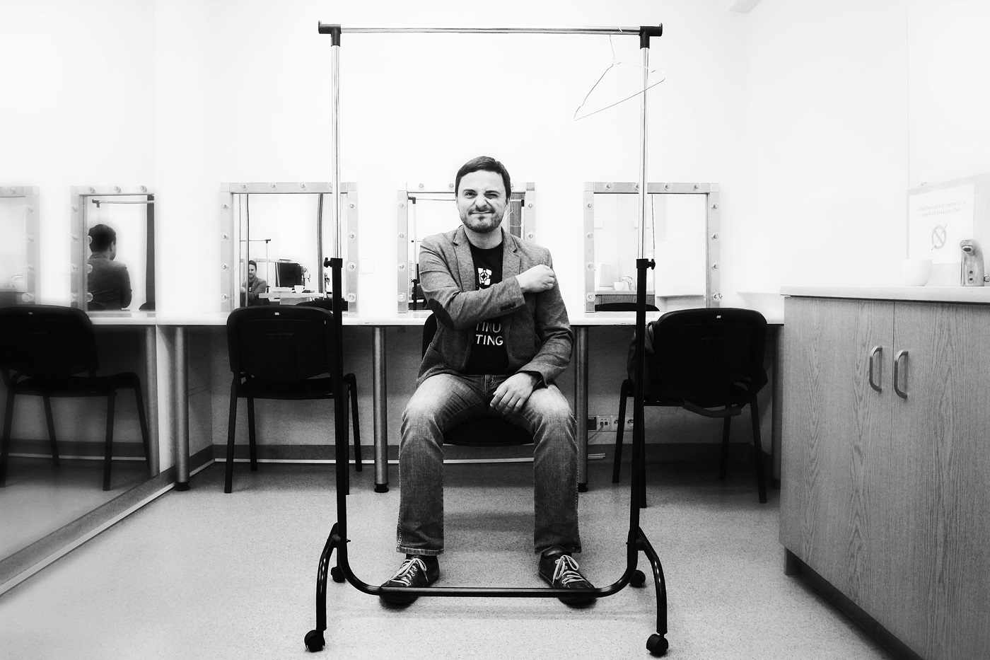 Czarno-białe zdjęcie mężczyzny siedzącego w garderobie. Za nim jest lada z trzema stanowiskami z krzesłami i lustrami otoczonymi lampkami. Mężczyzna siedzi na środkowym krześle, zwrócony plecami do lustra. Przed nim stoi wieszak na ubrania na kółkach i tworzy wokół mężczyzny symetryczną, prostokątną ramę.