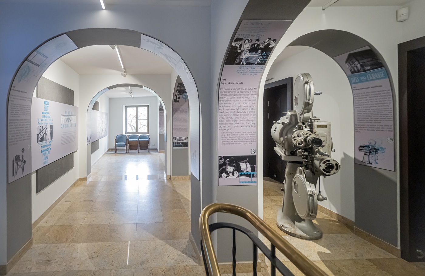 Część wystawy w Teatrze Starym poświęcona historii kina. Na pierwszym planie dwa łuki. W łuku po prawej stoi zabytkowy, wielki projektor filmowy na szpule z taśmą filmową.