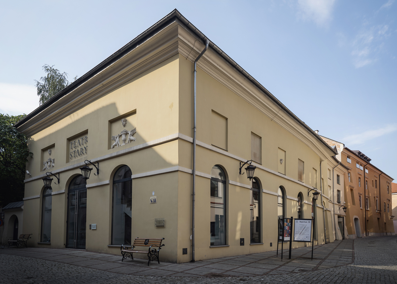 Zdjęcie budynku Teatru Starego w Lublinie. Ujęty jest róg budynku. Na prawo fasada od strony ulicy Jezuickiej. Na lewo boczna ściana od strony ulicy Dominikańskiej. Budynek jest bladożółty z białymi gzymsami na wysokości pierwszego piętra. Budynek ma wysokie okna z łukowym zakończeniem. Zaczynają się tuż nad ziemią i sięgają do pierwszego piętra. Nad nimi znajdują się na drugim piętrze płytkie wnęki. Wnęki od strony ulicy Dominikańskiej są puste. Od strony Jezuickiej są w nich płaskorzeźby, a w środkowej nad głównymi drzwiami jest napis "Teatr Stary". U góry nad budynkiem jest błękitne niebo.