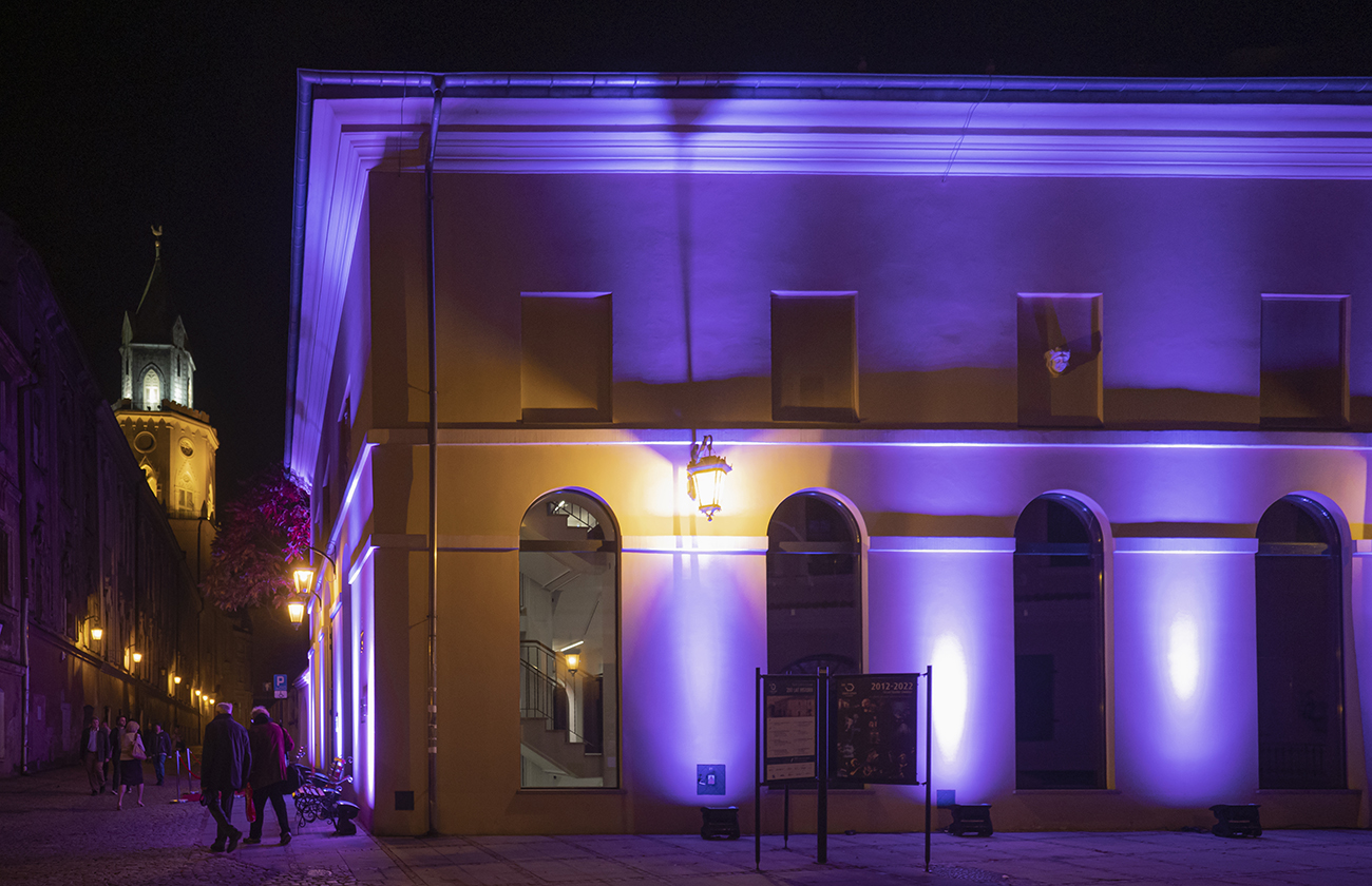 Zdjęcie dwupiętrowego budynku Teatru Starego w Lublinie nocą. Budynek jest oświetlony. W oświetleniu przeważa kolor jasnofioletowy. Rozproszony w jednym miejscu pomiędzy wysokimi okiennicami żółtym światłem latarni zamontowanej na ścianie.