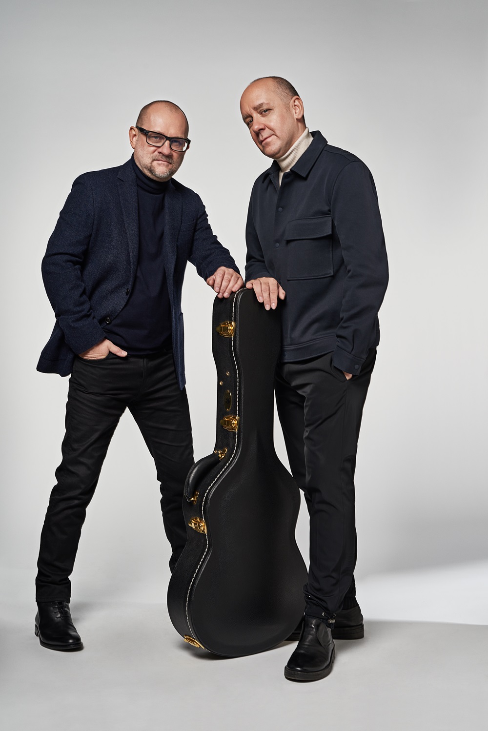 Zdjęcie dwóch mężczyzn w czarnych strojach. Pomiędzy nimi stoi czarny futerał na gitarę. Mężczyźni opierają po jednej ręce na futerale. Tło jest białe.