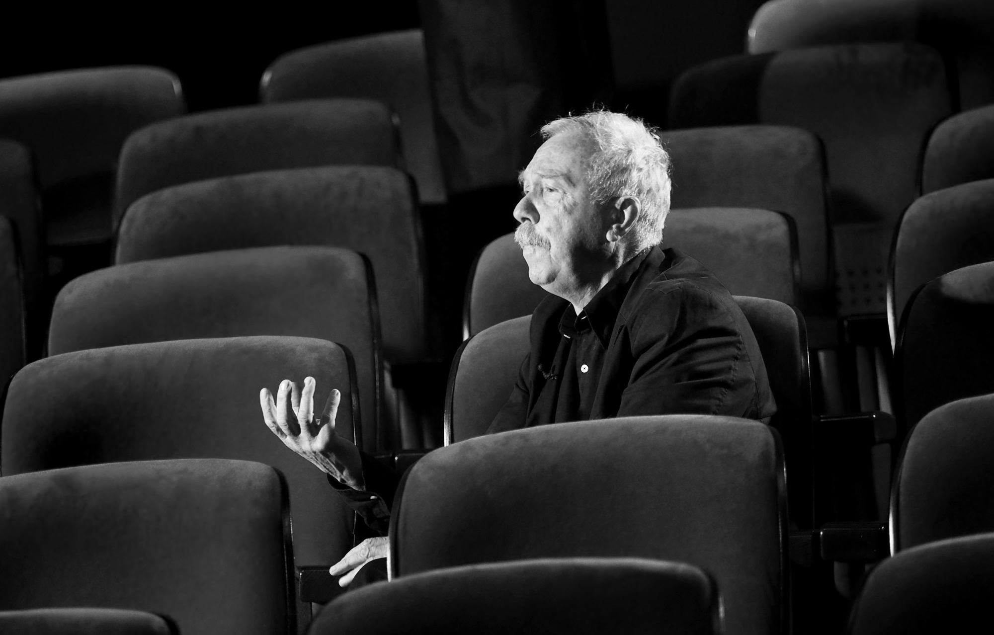Czarno-białe zdjęcie mężczyzny siedzącego na jednym z wielu foteli na widowni teatralnej. Mężczyzna ma jasne włosy i wąsy, jest ubrany na czarno. Odwraca głowę w lewo i gestykuluje. Fotele są ułożone w rzędy i zajmują całe tło zdjęcia.