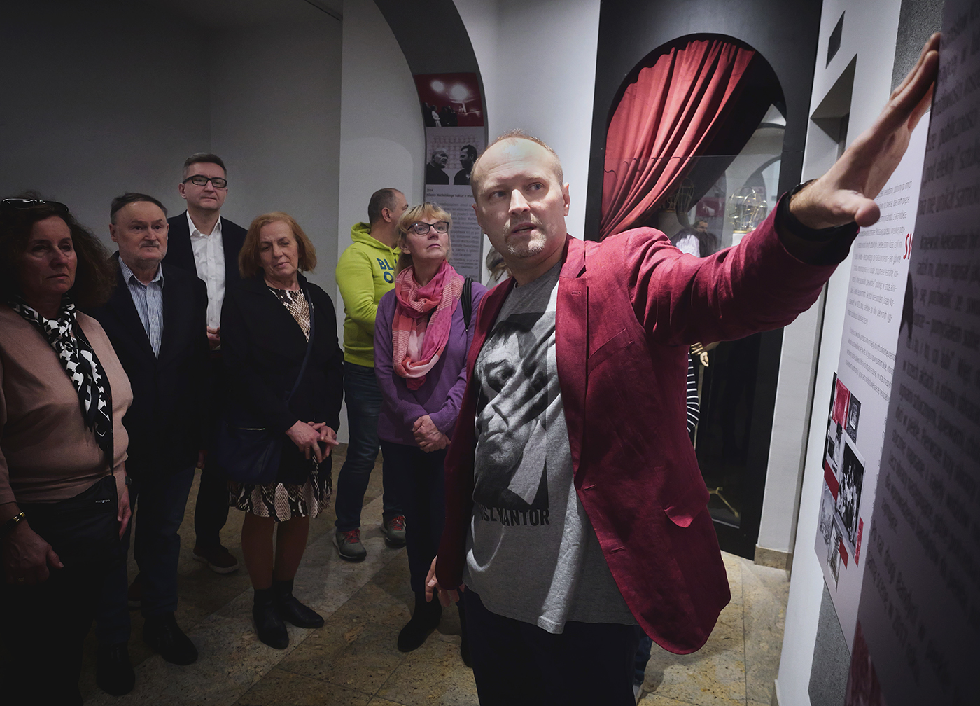 Zdjęcie przewodnika i grupy zwiedzających w Teatrze Starym. W przestrzeni w której znajdują się łuki. Wewnątrz łuków i na ścianach są tablice z tekstami i grafikami. W głębi po prawej stronie wnęka z uniesioną udrapowaną kotarą i eksponatami.