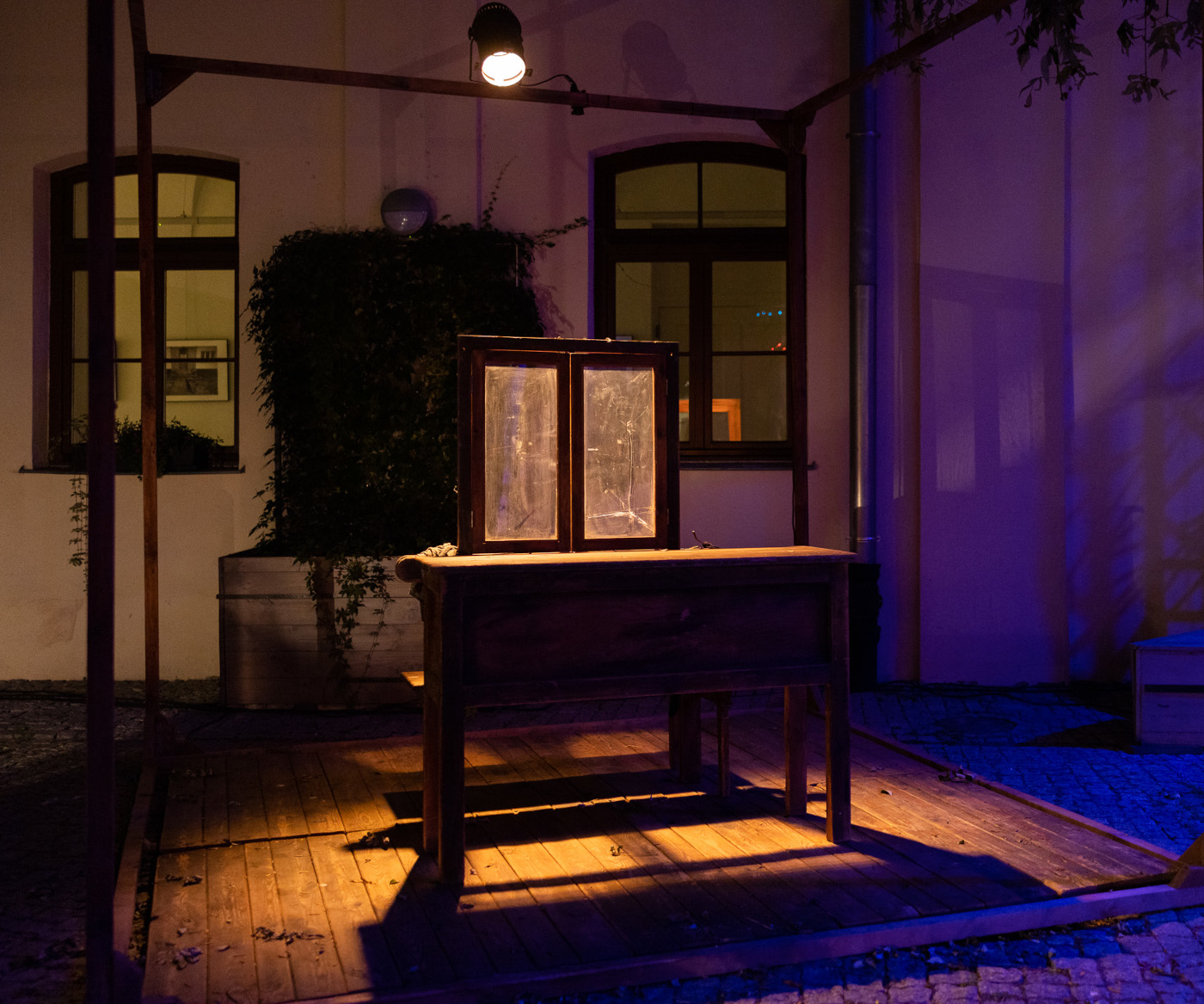 Fragment wystawy scenografii w plenerze po zmroku. Eksponowany jest podest na którym jest starodawna ławka szkolna, na niej stoi rama okienna z oknem z mętną szybą pękniętą w prawej części. Na te elementy pada ciepłe, żółtawe światło. W tle jest ściana jasnego budynku z dwoma oknami. Po prawej stronie jest oświetlona niebiskim światłem. Plamy światła płynnie się wtapiają, nie mają wyraźnych krawędzi.