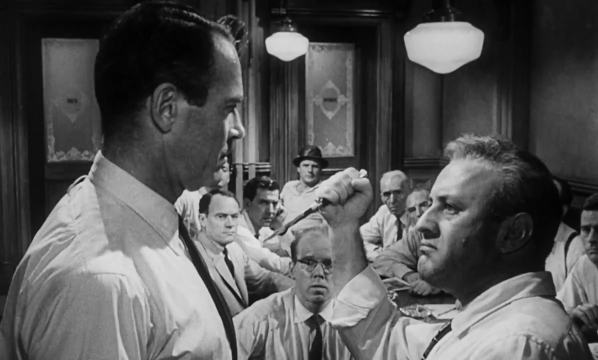 Czarno-biały kadr z filmu. Grupa mężczyzn w dużym pomieszczeniu. Na pierwszym planie dwóch mężczyzn stojących naprzeciwko siebie, jeden z nich trzyma nóż wymierzony w drugiego. Pozostali bacznie obserwują zajście. Wszyscy mężczyźni są ubrani w jasne koszule i ciemne, cienkie krawaty.