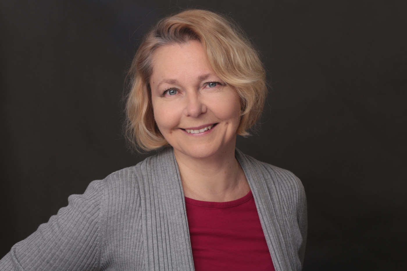 Zdjęcie portretowe uśmiechniętej, jasnowłosej kobiety w czerwonej bluzce i szarej narzutce. Tło jest ciemne.