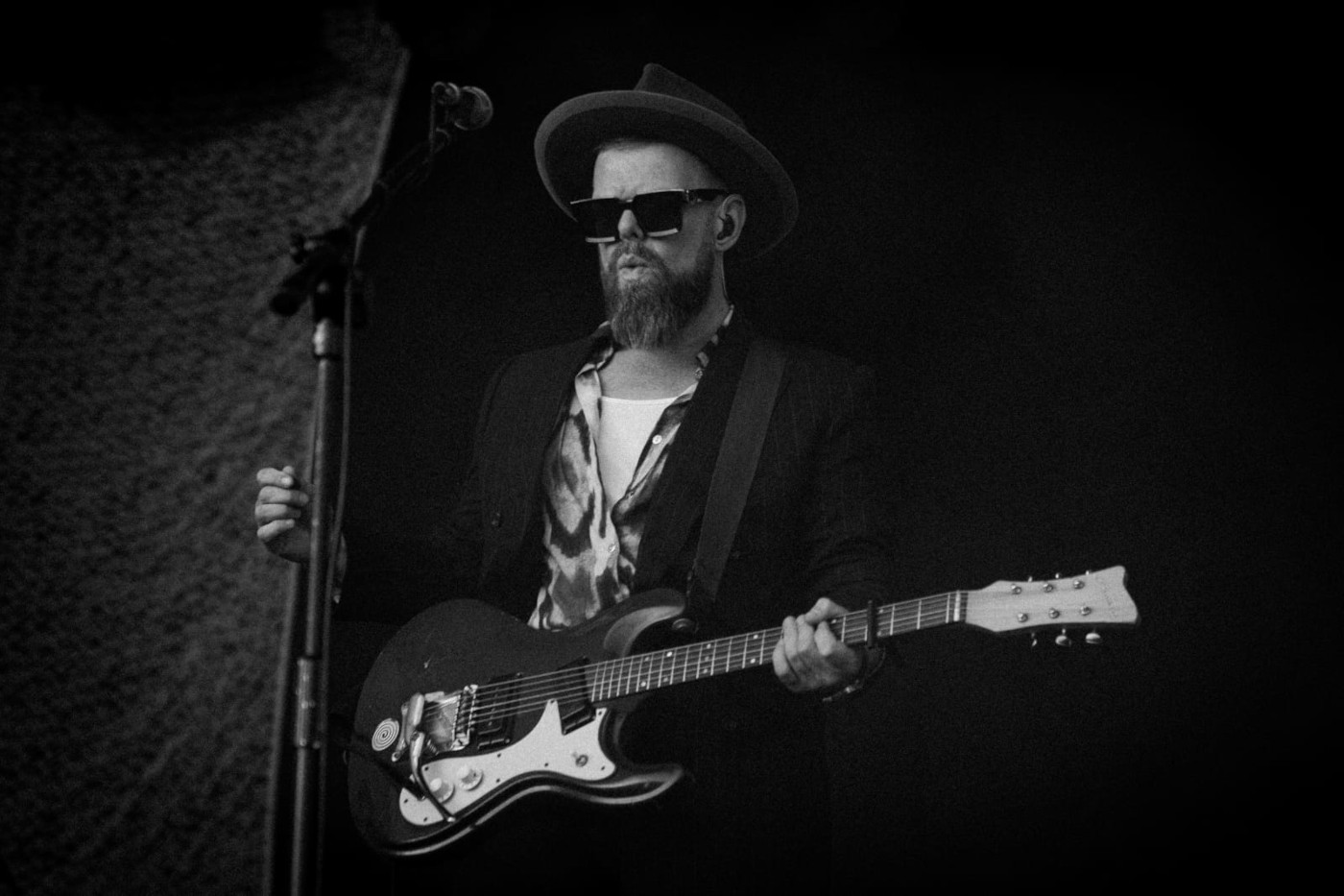 Czarno-białe zdjęcie Keva Foxa z gitarą elektryczną. Mężczyzna ma brodę, wąsy, ciemne okulary, kapelusz z okrągłym rondem, jest ubrany w jasną koszulkę z nadrukiem i ciemną marynarkę. Tło ciemne.